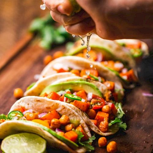 The image shows a hand squeezing lime over tacos filled with chickpeas, avocado, and vegetables, garnished with fresh herbs on a wooden board.