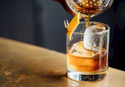A cocktail being poured into a glass with a large ice cube and a twist of citrus peel garnish, on a wooden surface.