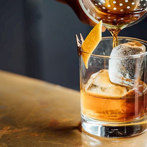A cocktail being poured into a glass with a large ice cube and a twist of citrus peel garnish, on a wooden surface.