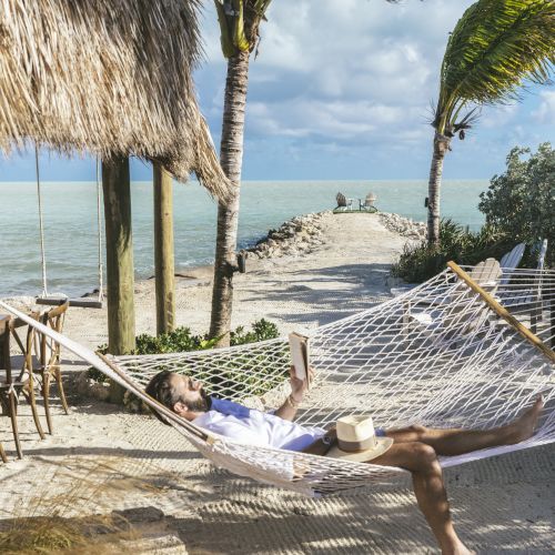 A person relaxes in a hammock by the beach, with palm trees swaying and the ocean in the background, enjoying a serene day.