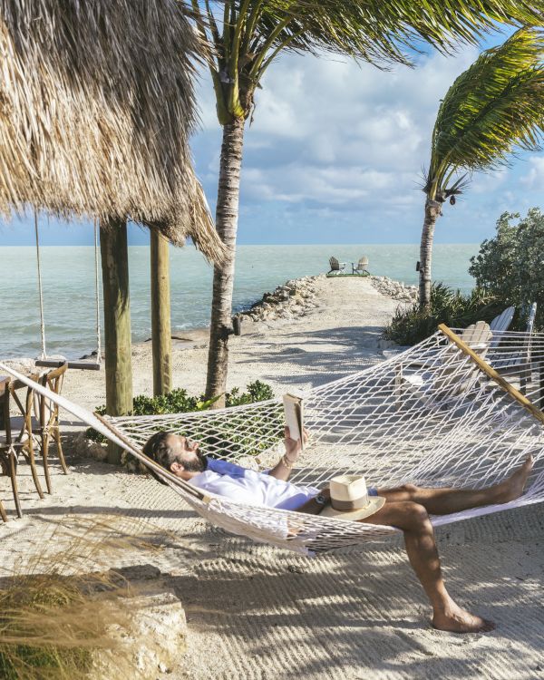 A person relaxes in a hammock by the beach, with palm trees swaying and the ocean in the background, enjoying a serene day.