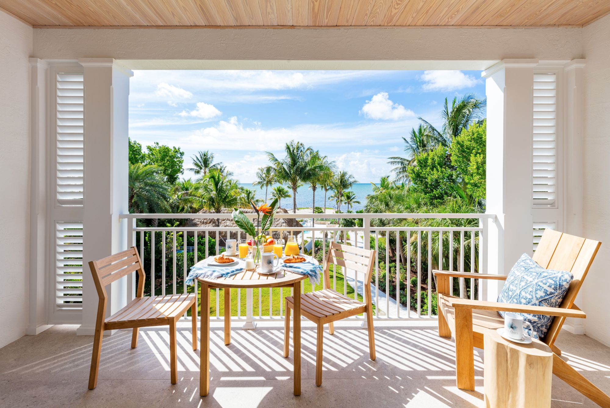 A balcony with a wooden table set, two chairs, and a side table. The view features palm trees, a garden, and the ocean with a sunny sky.