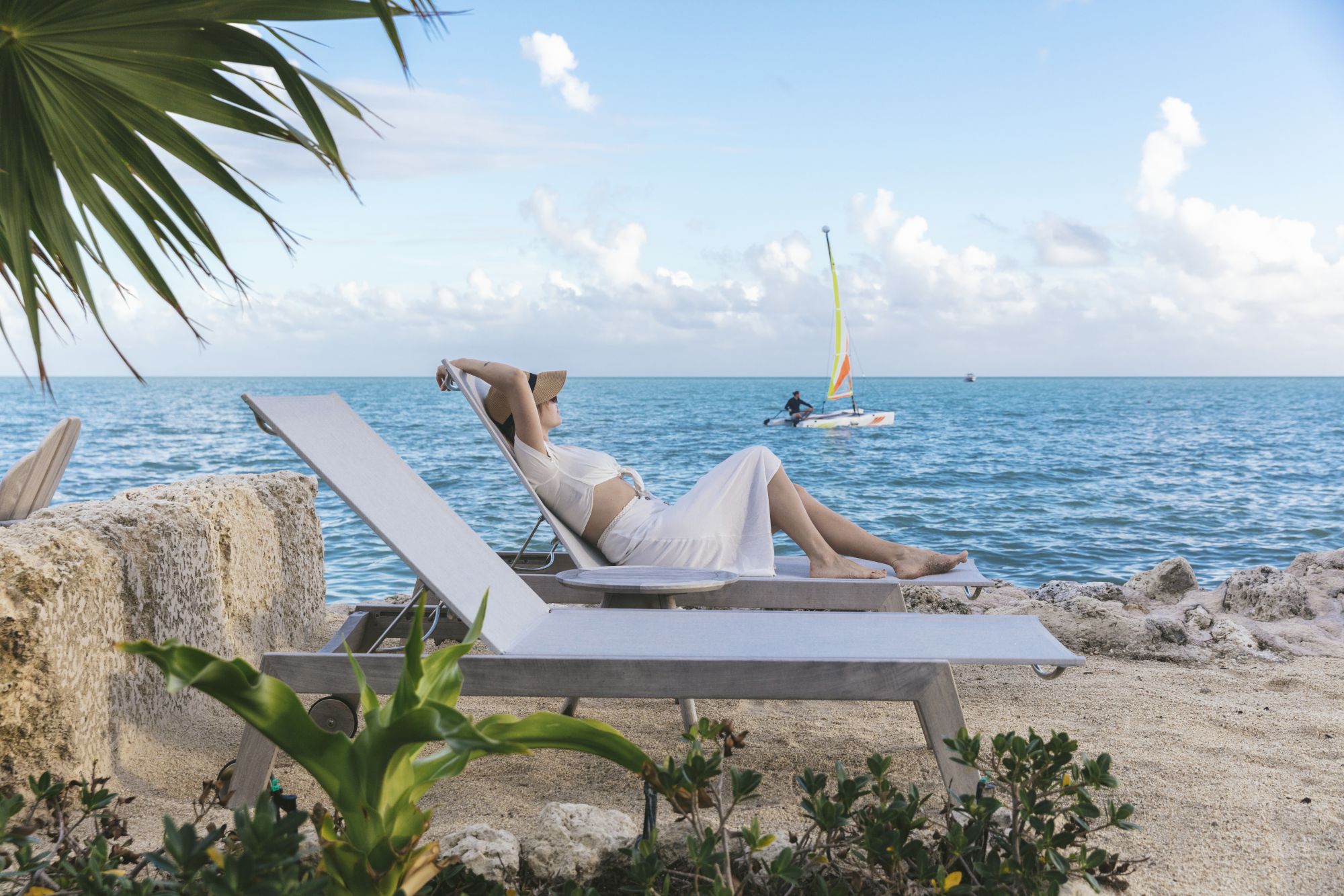 A person relaxes on a lounge chair by the sea, with a sailboat in the background, surrounded by greenery and a clear blue sky.