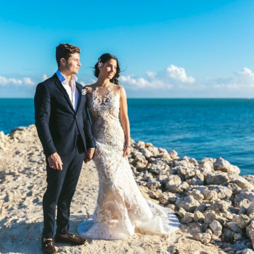 A couple in wedding attire stands on a rocky path by the ocean under a clear blue sky.