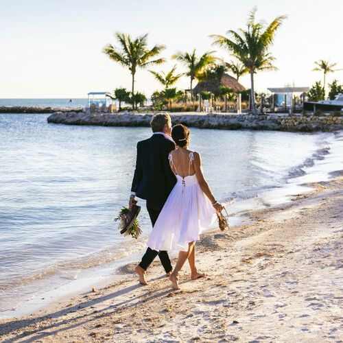 A couple, dressed in wedding attire, walks barefoot along a sandy beach near the water. Palm trees and clear skies are visible.