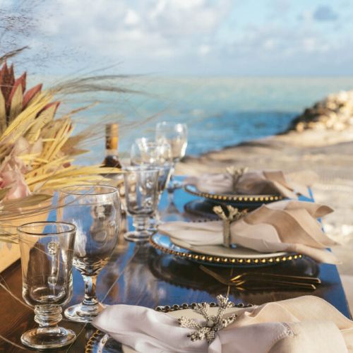 An elegantly set outdoor table with glassware and floral arrangements, overlooking the ocean on a sunny day.