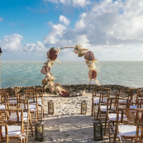 A beach wedding setup with an arch decorated with flowers, rows of chairs, and lanterns, overlooking the ocean with a partly cloudy sky.