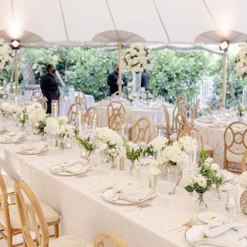 A beautifully set wedding or event table under a tent with white tablecloths, floral centerpieces, and elegant chairs, creating a serene setting.