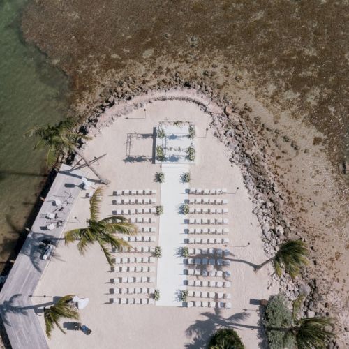 An aerial view of a beachfront wedding setup with rows of chairs facing a decorated arch, surrounded by palm trees and ocean water.