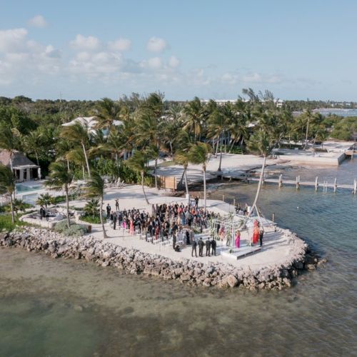 A wedding ceremony on a small beachfront surrounded by palm trees and water, with guests gathered around.