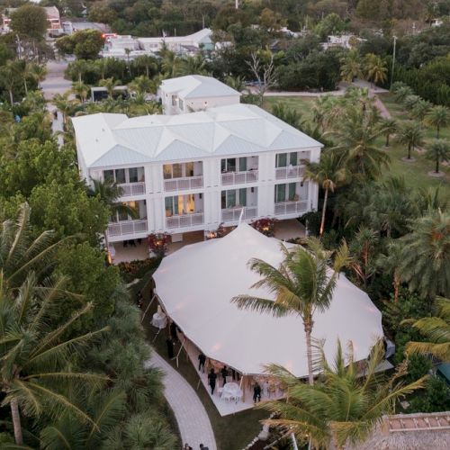 An aerial view of a white building with a tent outside, surrounded by palm trees and greenery.