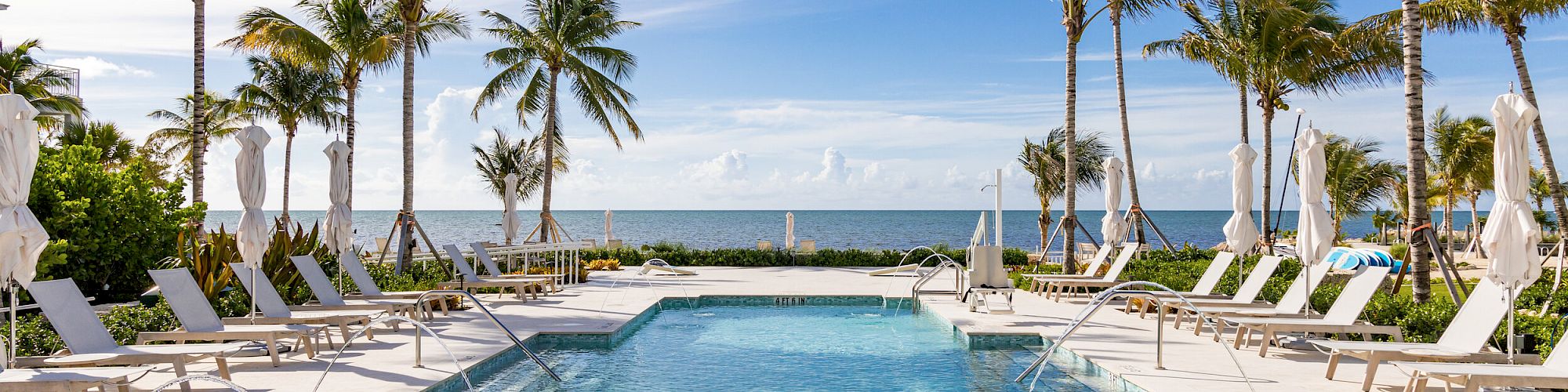 A luxurious poolside scene with lounge chairs, palm trees, and a beautiful view of the ocean under a blue sky, creating a tropical paradise setting.