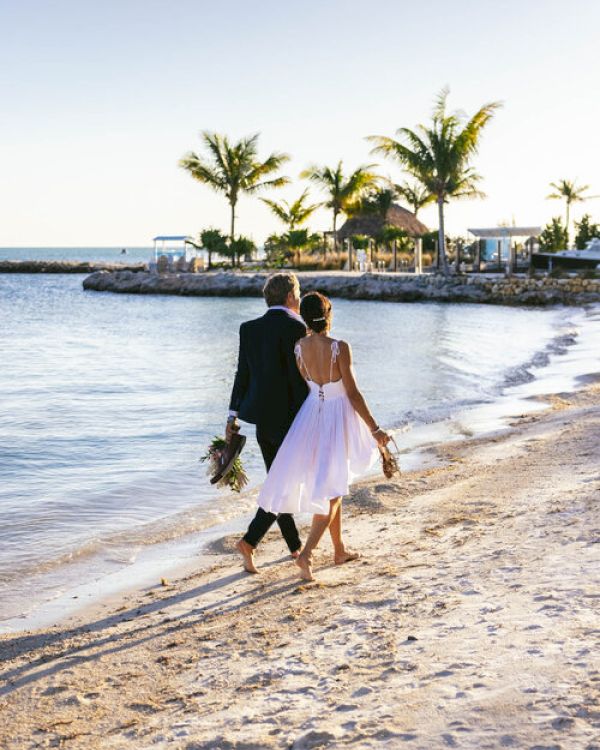 A couple, dressed formally, walks barefoot along a beach at sunset, holding hands and shoes, with palm trees and calm water in the background.