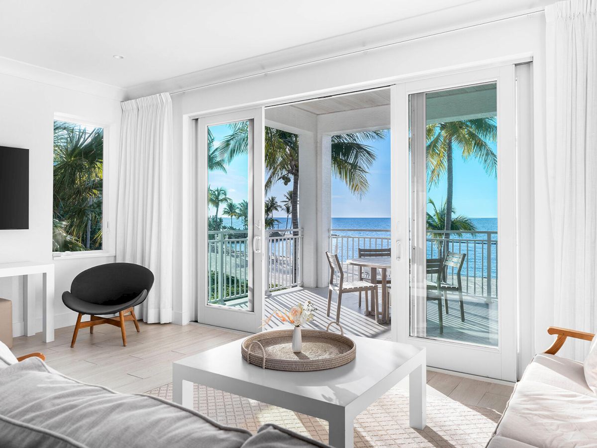 This image features a bright, modern living room with white decor, a large TV, and a balcony view of the ocean and palm trees.