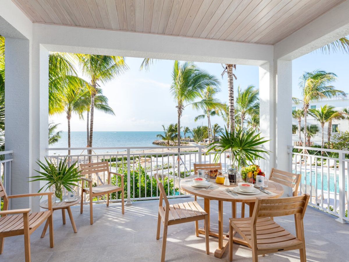 A tropical patio with wooden furniture overlooks a pool, palm trees, and the ocean. Breakfast is set on the table, surrounded by lush greenery.