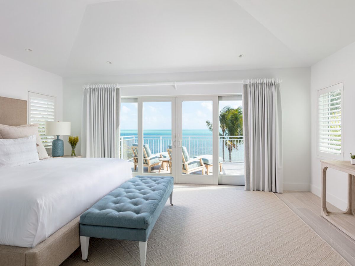 A bright bedroom with a beige bed and blue bench faces glass doors opening to a balcony with ocean views. A TV and side table adorn the right wall.