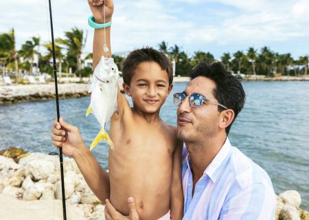 A child proudly holds up a fish they caught while an adult supports them. They are by the water, with a fishing rod and a clear sky in the background.