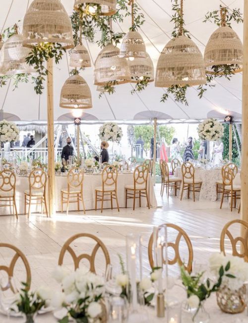 The image shows a beautifully decorated wedding tent with elegant chairs, floral arrangements, and hanging lights, prepared for a celebration.