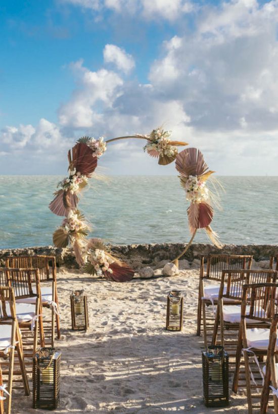 A beach wedding setup with wooden chairs arranged in rows facing an arch decorated with flowers and leaves, and a view of the ocean under a partly cloudy sky.