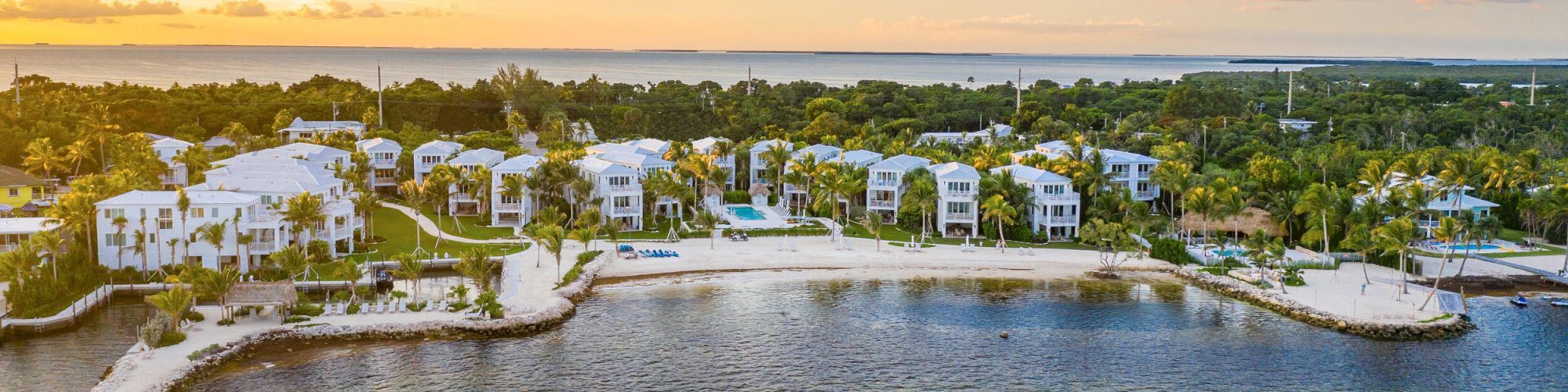 An aerial view of a coastal resort with white buildings, a beach, and lush greenery at sunset, captured along a serene waterfront.