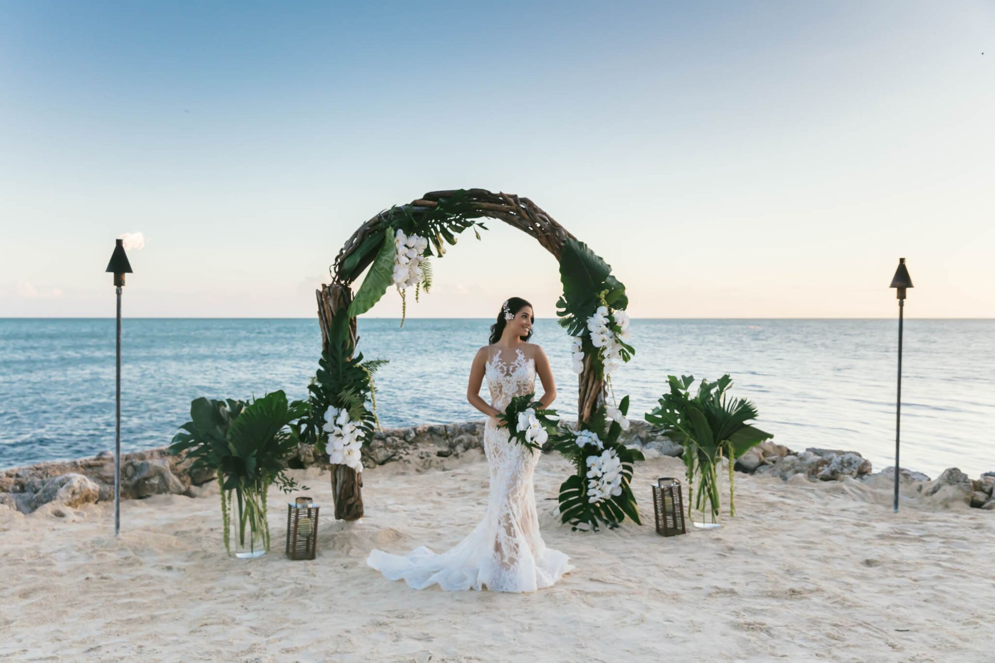 A bride stands in front of a floral arch on a beach, with the ocean in the background and tiki torches on either side, creating a serene scene.