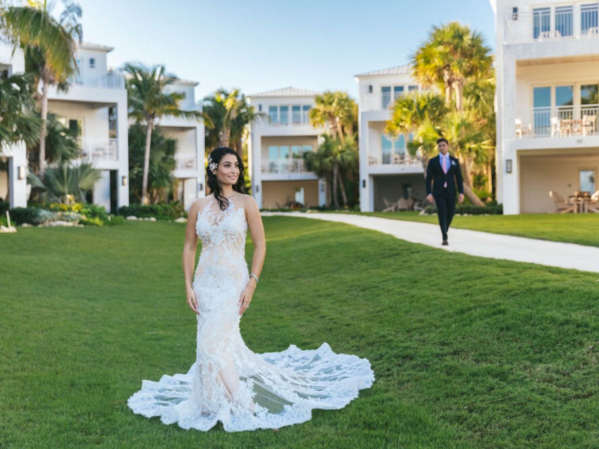 A woman in a wedding dress stands on a lawn with a bouquet, while a man in a suit walks toward her. There are buildings and palm trees in the background.