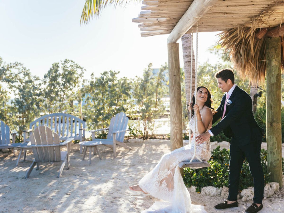 A couple, with the bride on a swing and groom behind her, poses in a scenic outdoor setting with beach chairs and greenery.