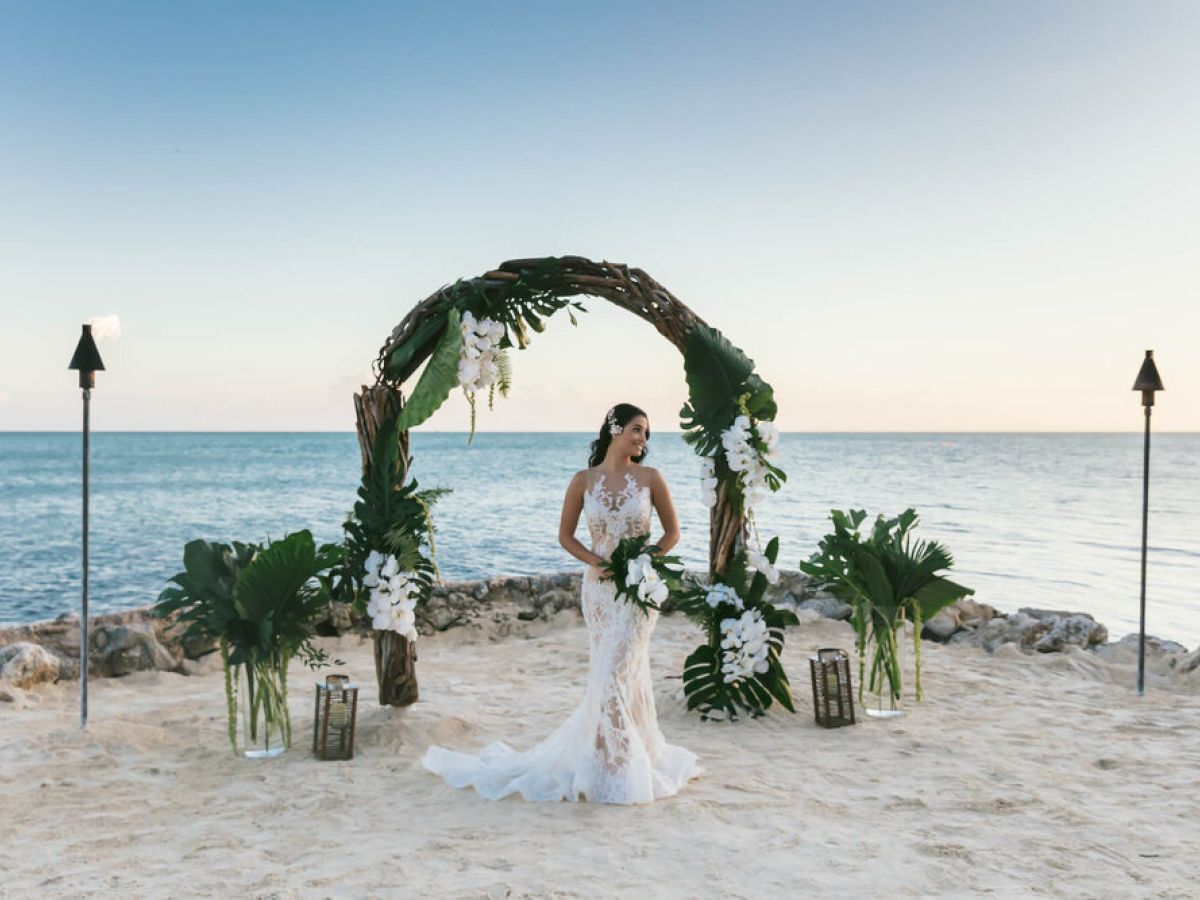 A bride in a white dress stands under a floral arch on a sandy beach, with the ocean in the background and tiki torches on either side.