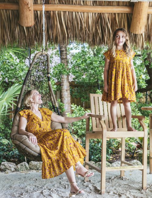 An adult and a child, both wearing yellow dresses, are in an outdoor setting with tropical plants. The adult is seated, and the child stands on a chair.