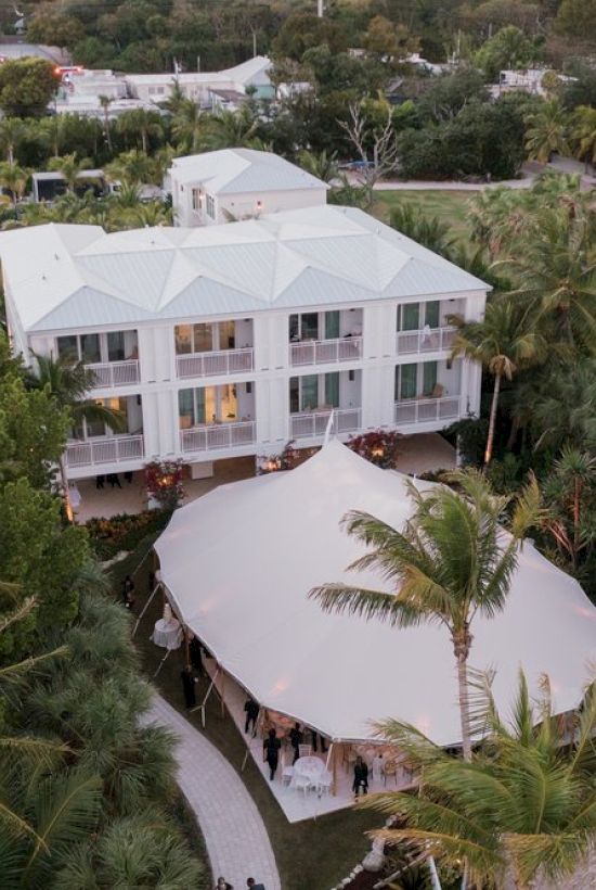 Aerial view of a tropical property with a white tent, surrounded by palm trees and a multi-story building in the background, with green lawns.