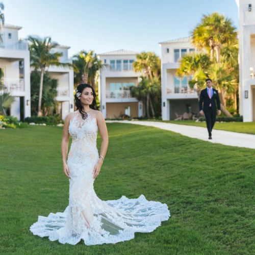 A woman in a wedding dress stands on a green lawn with buildings in the background as a man in a suit walks towards her.