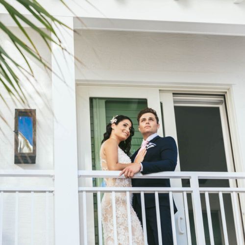 A couple is standing on a balcony, with the woman in a white dress and the man in a suit, both looking off into the distance, framed by white walls.