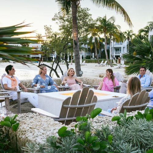 A group of people sit in a tropical outdoor setting with palm trees, wearing casual attire, engaged in conversation around a fire pit.