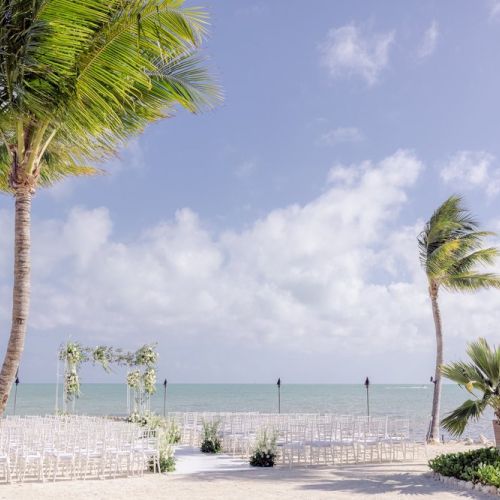 Beach wedding setup with chairs, flower arch, and palm trees under a clear, partly cloudy sky by the ocean, creating a serene atmosphere.