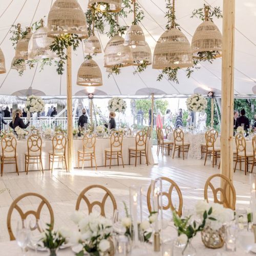 The image shows a decorated event tent with numerous round tables and wooden chairs, hanging wicker lights, and floral arrangements.