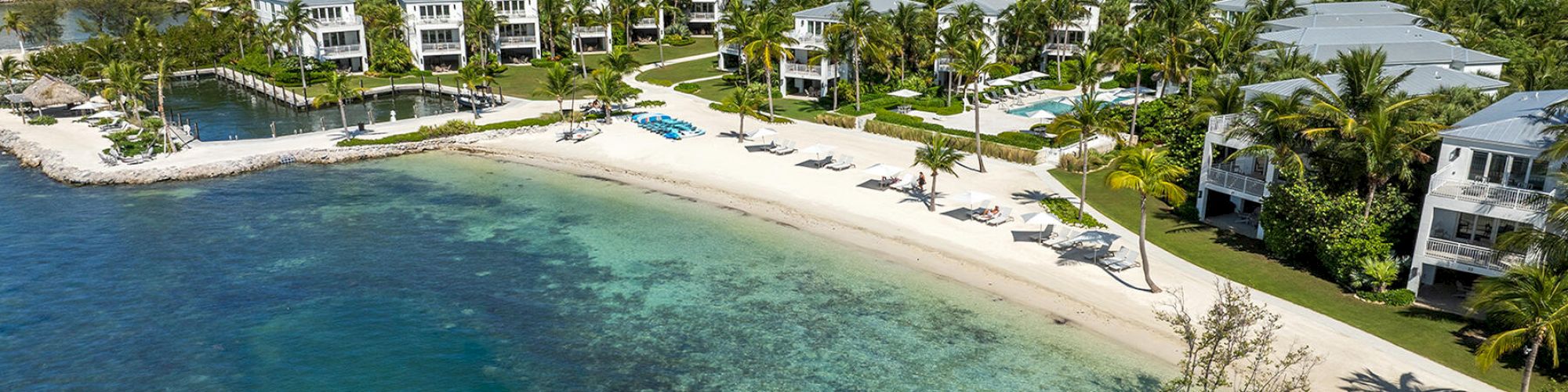 Aerial view of a beachfront resort with white sandy beach, clear turquoise water, and multiple buildings surrounded by lush greenery and palm trees.