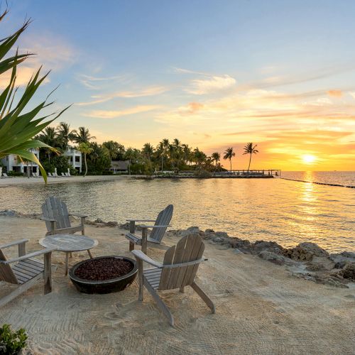 A peaceful beach scene at sunset with wooden chairs around a fire pit, overlooking the water and distant palm trees.