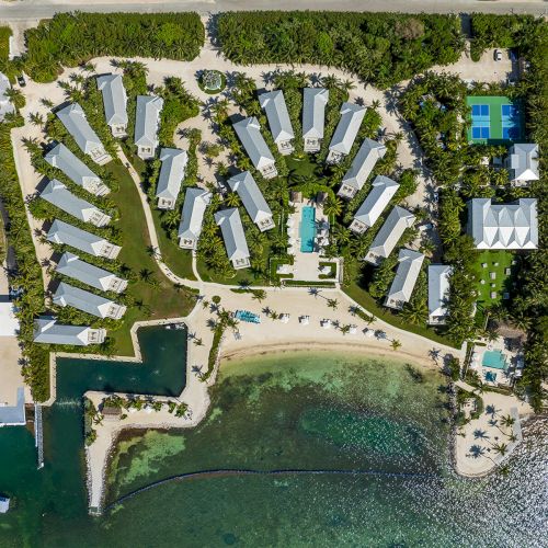 An aerial view of a beachside resort with multiple buildings, a pool, dock, tennis courts, and lush greenery by the sea, ending the sentence.