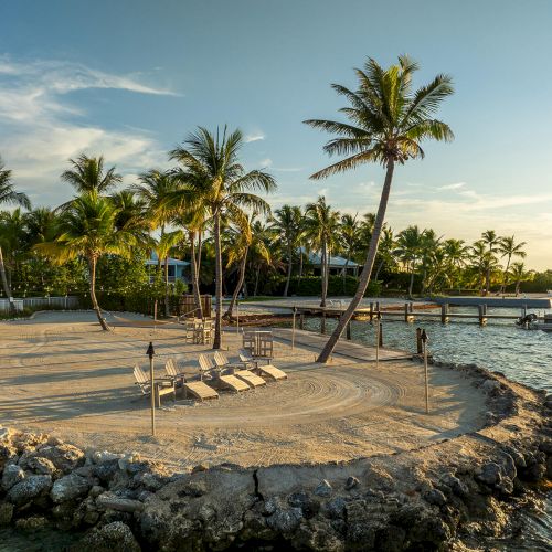 A tropical island setting with palm trees, lounge chairs, rocks, a dock, and a boat on the water during sunset or sunrise.