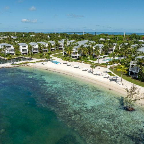 A scenic beachfront with clear water, various buildings, green lawns, and palm trees lining the shore under a bright blue sky.