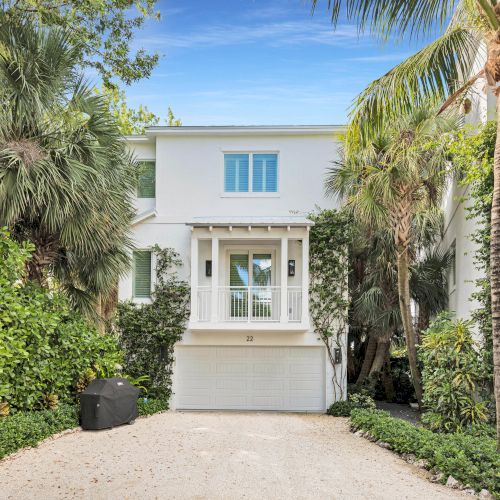 A white two-story house surrounded by lush greenery and palm trees, with a gravel driveway leading to the garage, under a clear blue sky.