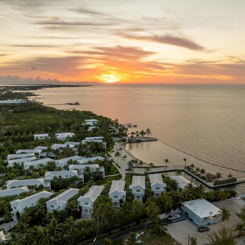 An aerial view of a coastal area at sunset, featuring houses, palm trees, and calm waters meeting the horizon.