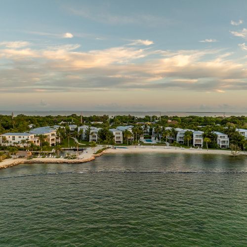 The image shows a coastal residential area with waterfront properties surrounded by trees and a calm body of water under a partly cloudy sky.