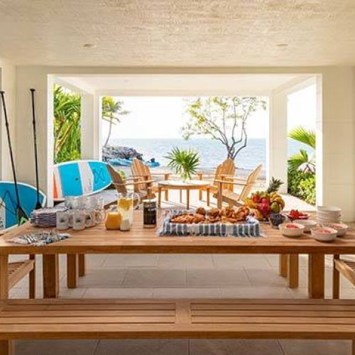 A beachfront patio features a wooden table with breakfast items, surrounded by chairs, surfboards, and tropical greenery overlooking the sea.