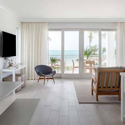 A modern living room with a coastal view, featuring white decor, a wall-mounted TV, furniture, and a patio leading to the beach.