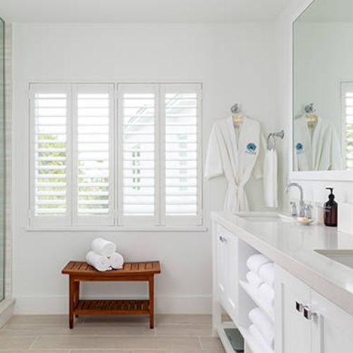 A modern bathroom with a glass shower, two sinks, towels, robes, and a wooden bench near a window. Neat and minimalistic design.