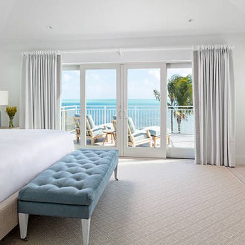 A bright bedroom with a large bed, blue bench, and ocean view through glass doors, leading to a balcony with chairs outside.