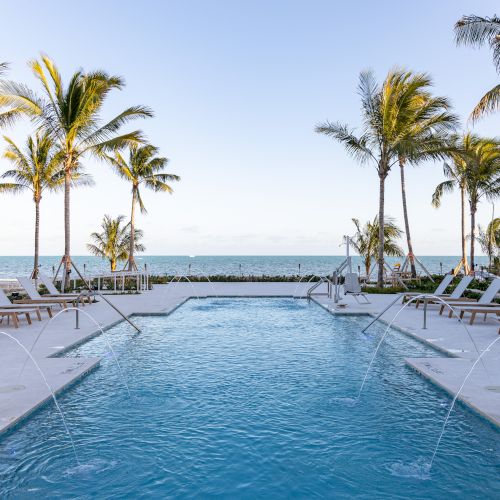 A pool with fountains surrounded by lounge chairs and palm trees, overlooking the ocean under a clear blue sky.
