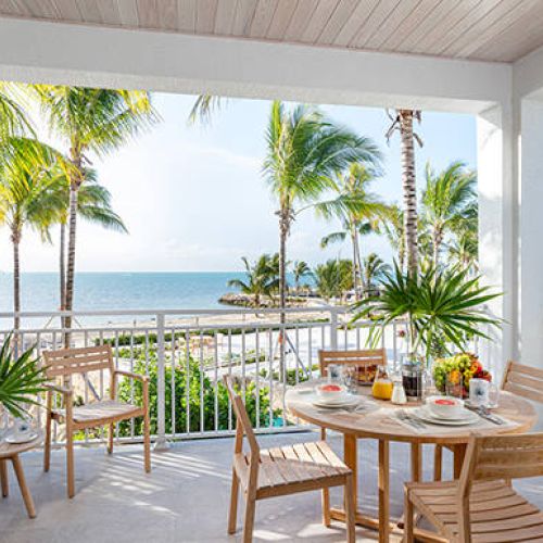 A tropical balcony with wooden furniture overlooks the ocean, featuring palm trees and a pool area. A set table suggests a meal setting.