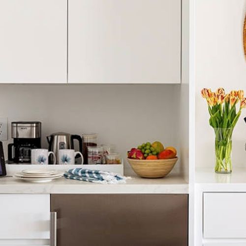 A modern kitchen counter with a tablet, coffee maker, mugs, fruit bowl, flowers, a wine bottle in an ice bucket, and two wine glasses.